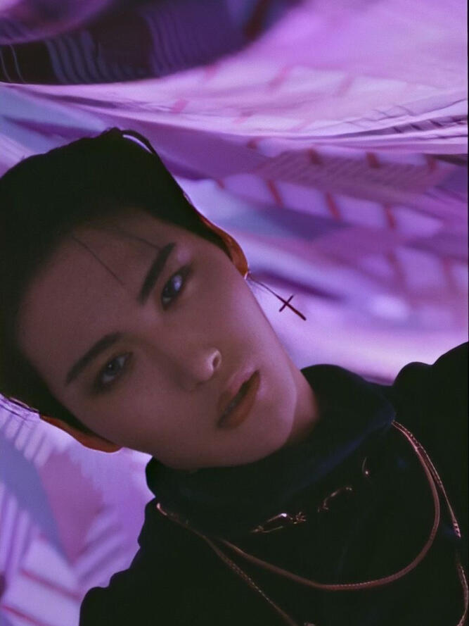 A picture from the music video of “Illusion” by the K-pop group Ateez showing Park Seonghwa. In this photo one can see Seonghwa’s face and shoulders. He wears black clothes, has short black hair and stares into the camera. The background is light purple.